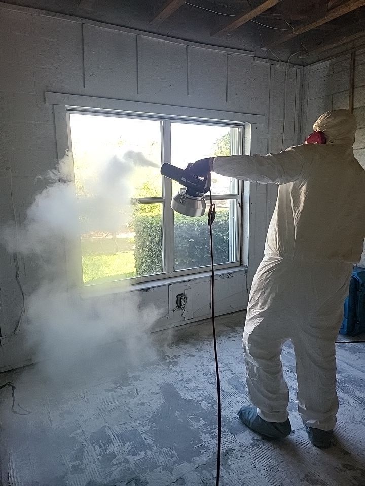 Technician in full PPE thermal fogging stripped block wall room near window